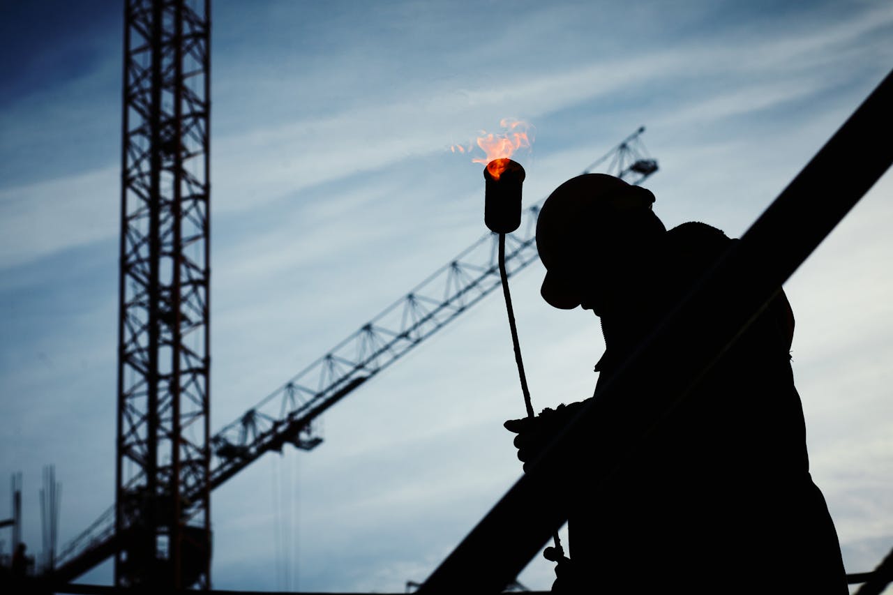 Silhouette of a construction worker using a blowtorch at a building site against a crane-filled skyline.